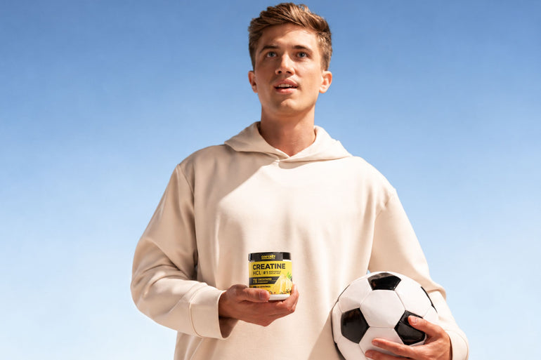 A young man in a beige hoodie holds a jar of CON-CRĒT Creatine HCl powder and a soccer ball against a clear blue sky background.
