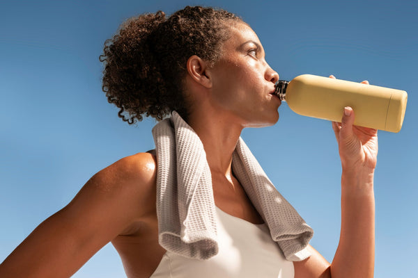 A woman outdoors drinking from a matte yellow reusable water bottle, with a white tank top and a gray towel around her neck against a clear blue sky.